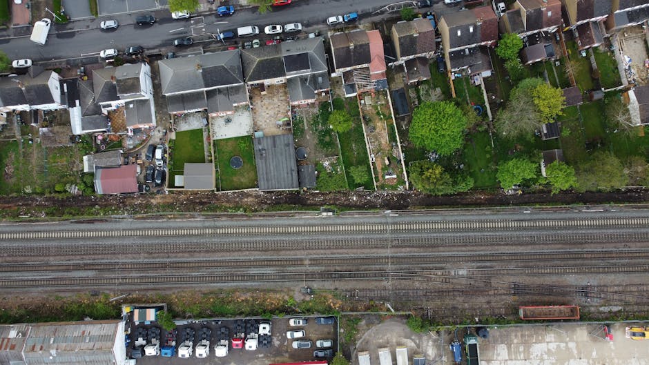 An aerial view of a row of residential garden plots situated behind terraced houses along a street, showcasing varied landscaping and outdoor maintenance. The gardens feature lush green lawns, some wi