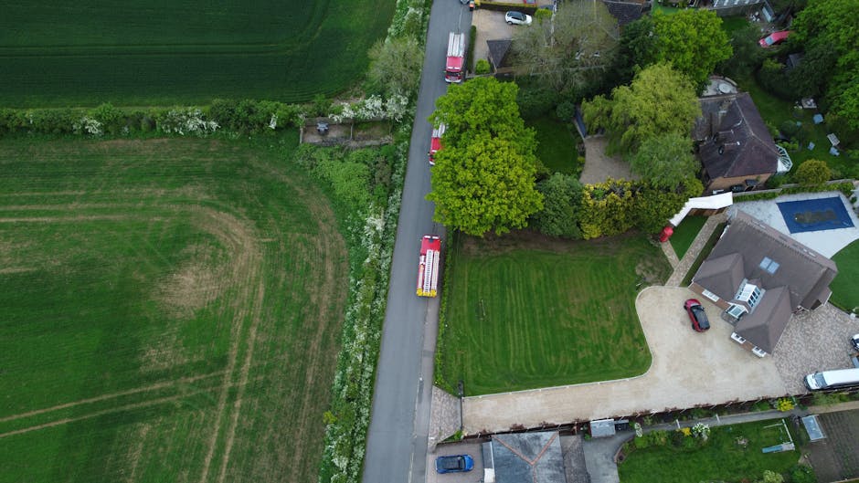 An aerial view of a row of residential garden plots situated behind terraced houses along a street, showcasing varied landscaping and outdoor maintenance. The gardens feature lush green lawns, some wi